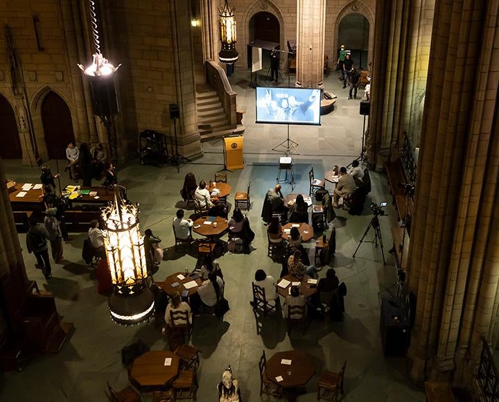 People sit around tables in the Cathedral Commons Room. A screen and podium are at the front of the room.