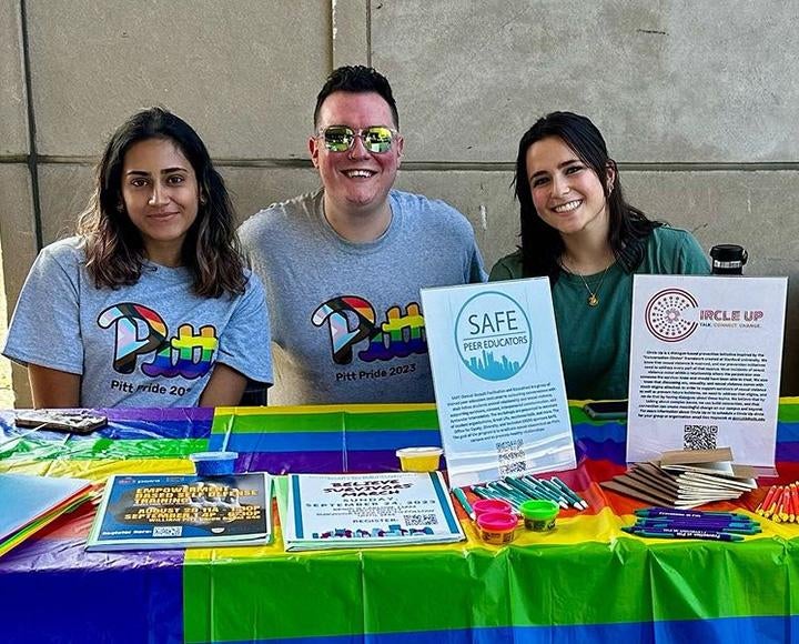 Three people sit behind a rainbow table with flyers and prizes