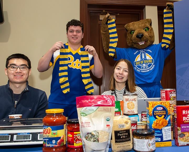 Two people sit in front of a table of food donations, while a person and a mascot hold up socks behind them