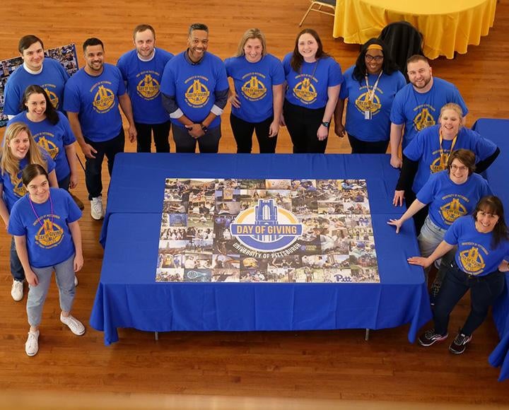 Volunteers in blue shirts stand around a completed Pitt Day of Giving puzzle