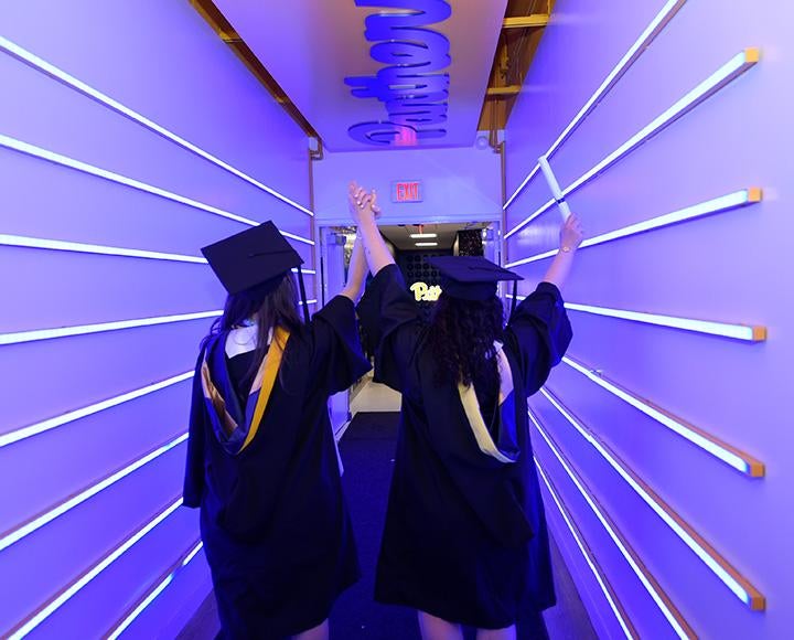 Two graduates raise held hands as they walk down a blue hallway