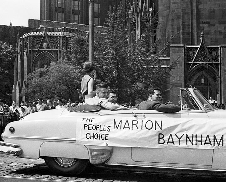 A black and white photo of a car in a parade. A banner on the said reads The Peoples Choice Marion Baynham