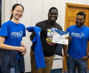 A person holds a certificate and T-shirt while standing in between two people in the same blue T-shirt that reads RNEL