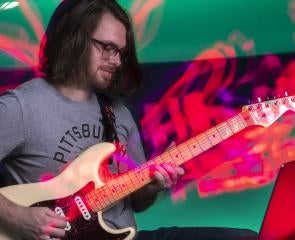A person looks at a laptop while playing guitar surrounded by neon illustrations