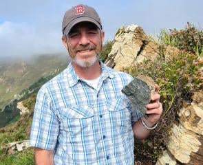 Kerrigan holds up a rock. A foggy seaside cliff is behind him.