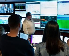 Four students watch as an instructor points at a wall of screens displaying data