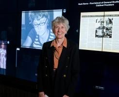 An older woman stands in front of a photo of her younger self on a screen