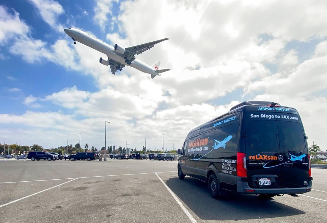 A plane flies low overhead a black van in an LAX parking lot