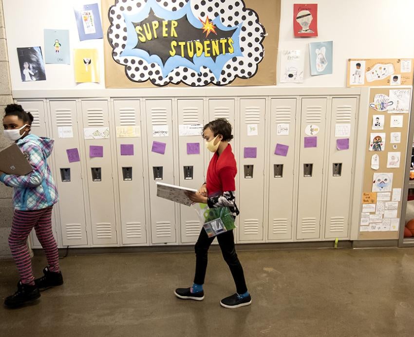 Two students with masks walking through a hallway with lockers, a poster with "Super Students" hangs on the wall.