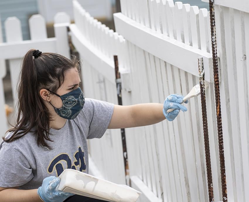 Pitt student with face mask painting white fence
