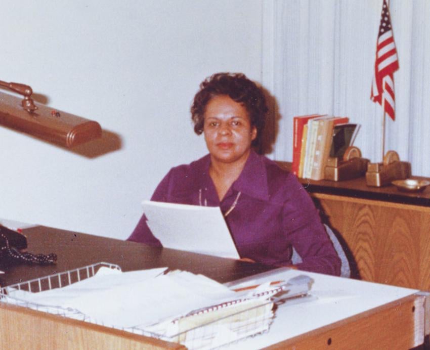 Gertrude Wade in a purple dress shirt sitting at a desk.