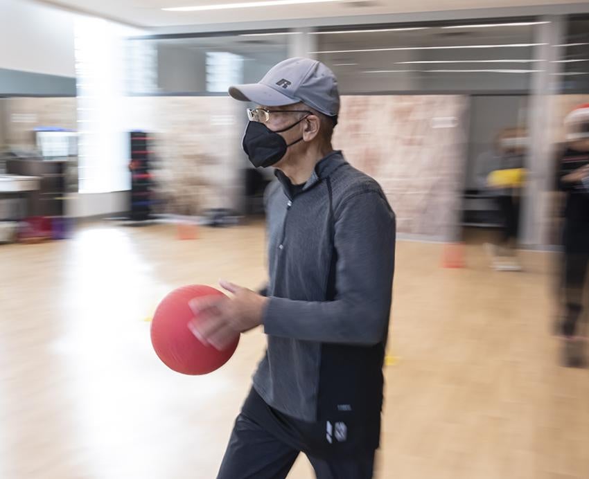 Man with grey shirt and hat holding a red ball in exercise room 