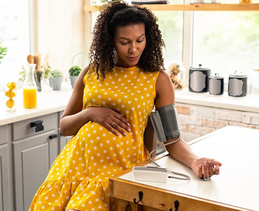 Pregnant woman in yellow dress sitting in a kitchen using a blood pressure monitor.