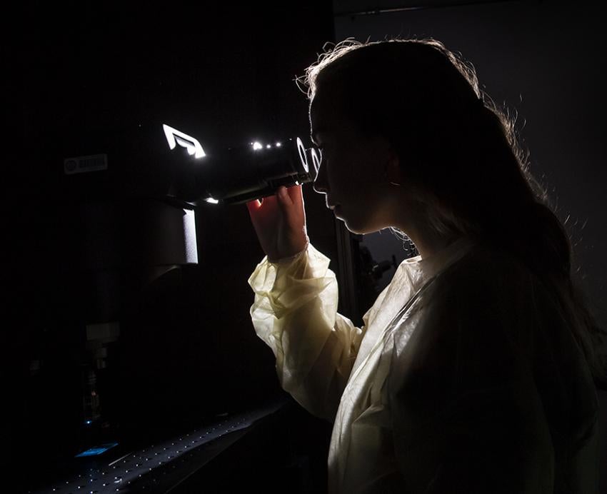 Woman looking through microscope in a dark room with computer in background