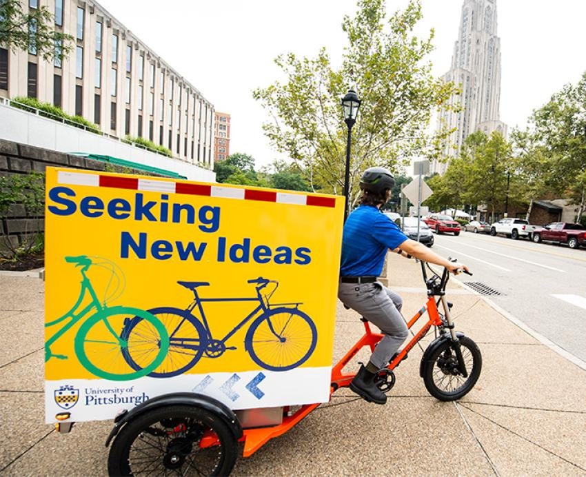 A person rides a bike that is carrying a yellow sign that reads Seeking New Ideas.