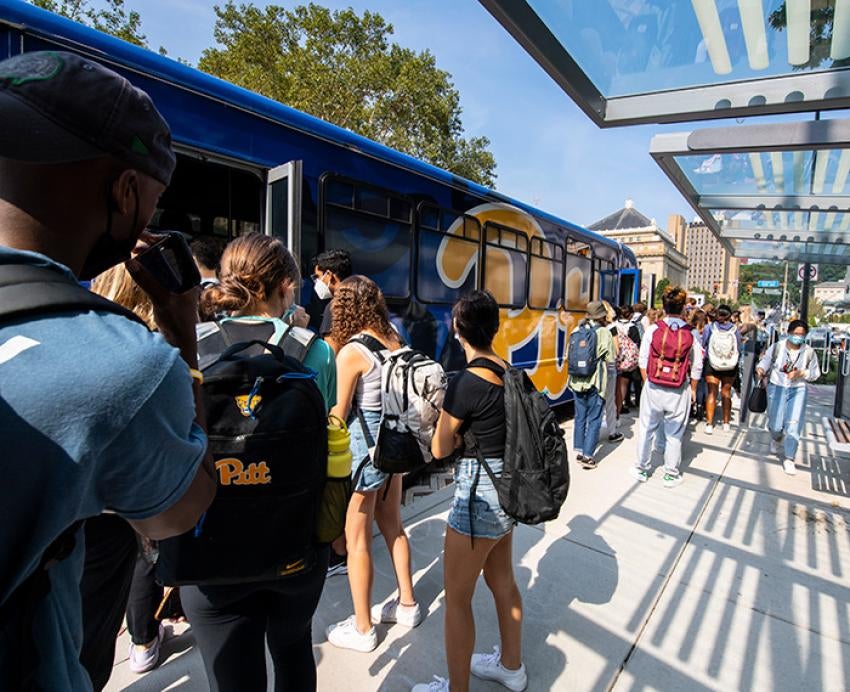 A crowd of people board a blue shuttle bus with a Pitt script logo
