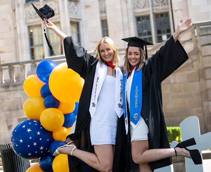 Two people in white dresses and graduation gowns pose in front of blue and yellow balloons