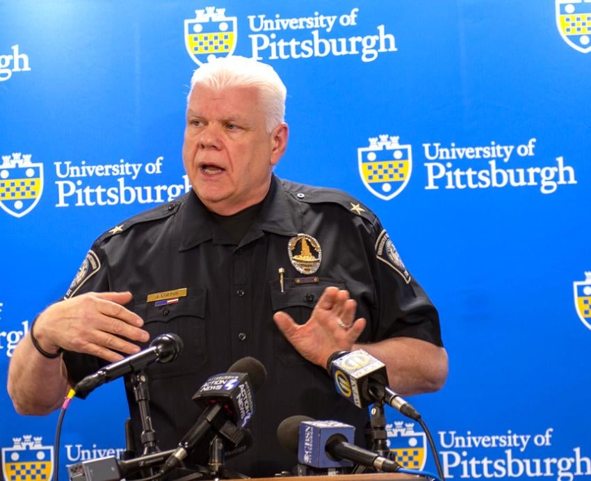 James Loftus speaks into microphones at a podium in a black Pitt Police uniform