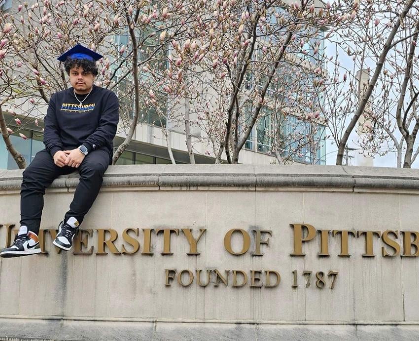 Knight sits on the marble University of Pittsburgh sign wearing all black and a blue graduation cap