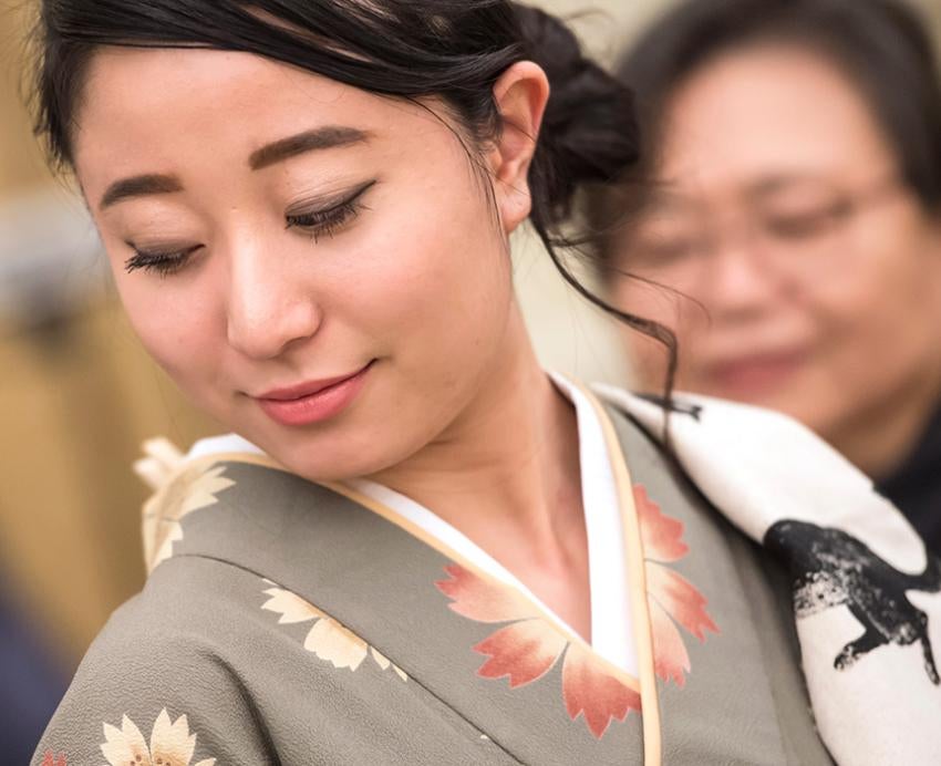Two people stand behind a girl in a kimono. Off-camera, they are tying a ceremonial sash.