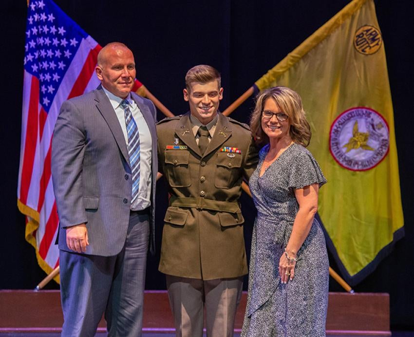 Erickson stands between his parents in an Army uniform. A U.S. flag and a yellow R.O.T.C. flag hang in the background.