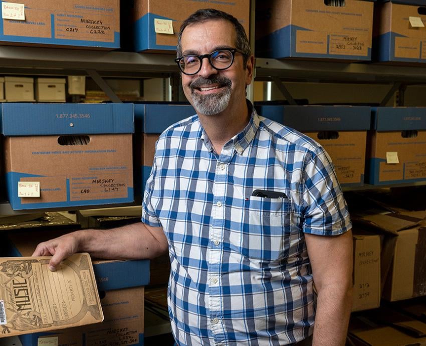 Cassaro holds an antique music book in front of a shelf of boxes of archives
