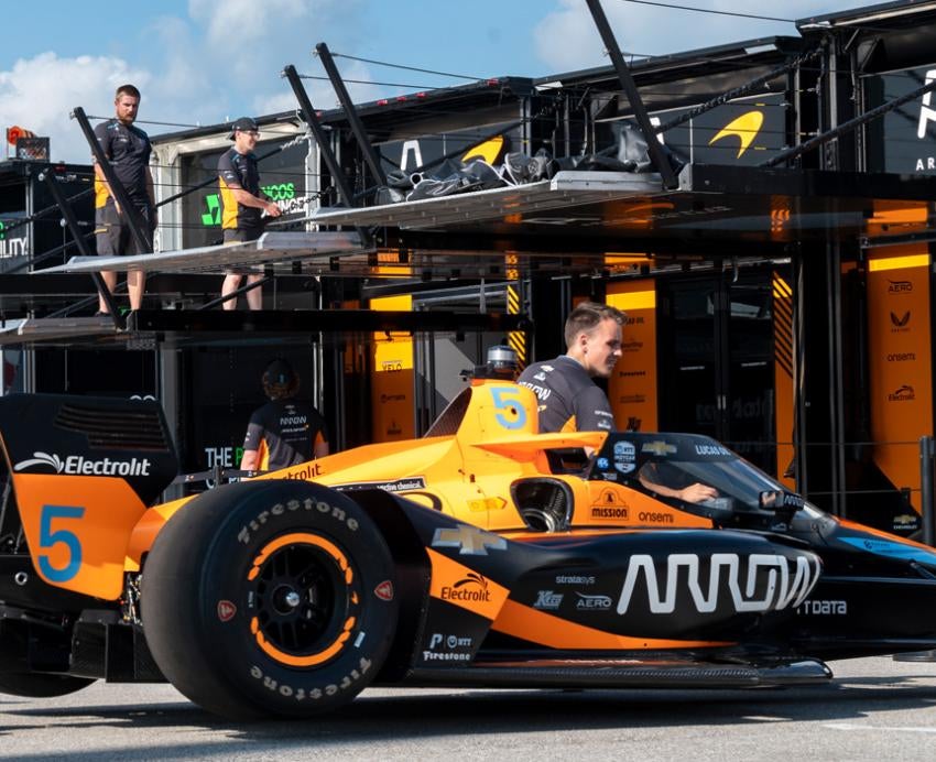 Team members pull an orange and black racing car from its garage onto the track