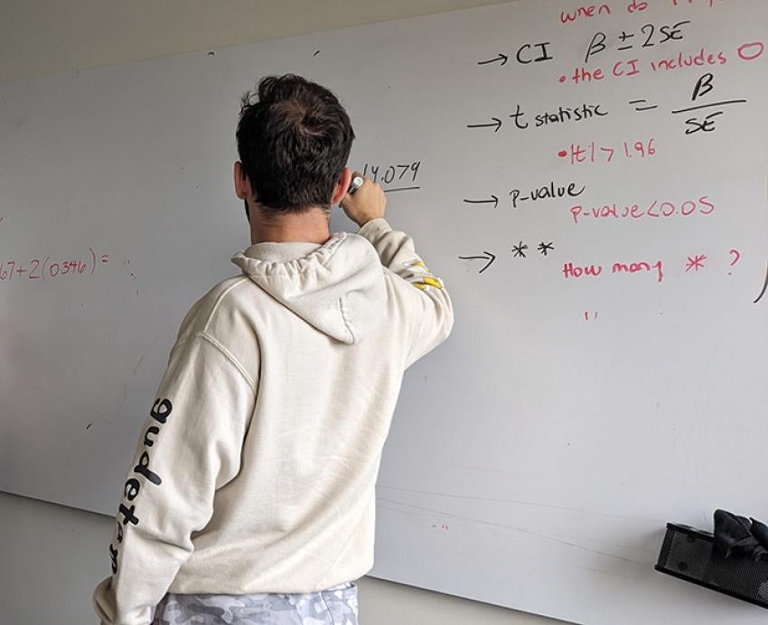 A student in a beige hoodie writes on a dry erase board