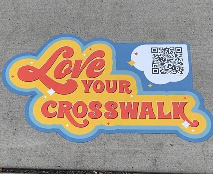 a sticker on the ground that says Love Your Crosswalk and has a QR code