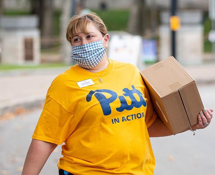 A masked person wearing a Pitt in Action shirt carries a cardboard box.