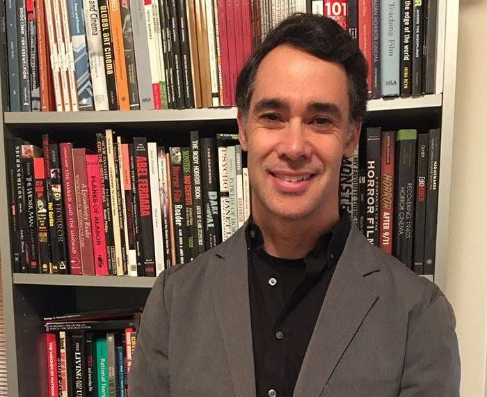 Adam Lowenstein in a grey suit and black shirt in front of a bookshelf