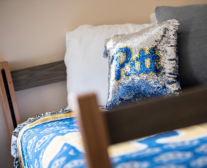 A sequined Pitt pillow and throw blanket on a residence hall bed