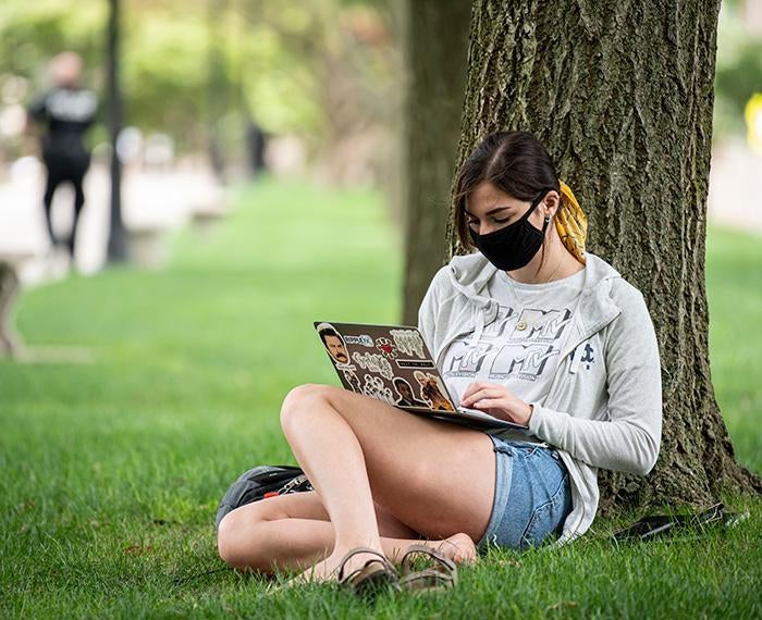 A masked person sits against a tree with a laptop
