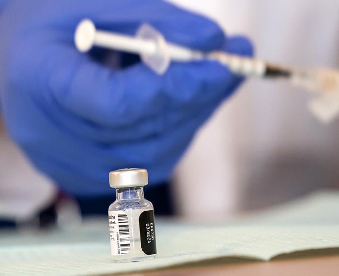 A vaccine bottle on a table with a blue gloved hand holding a syringe in the background