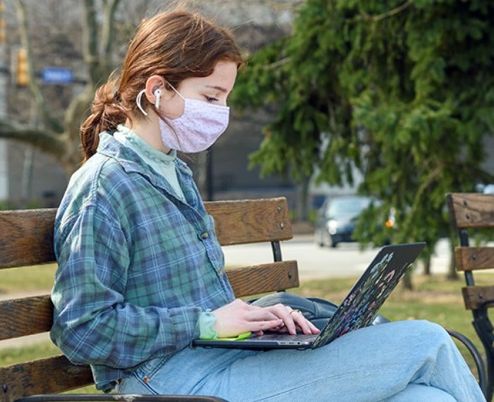 A person in a plaid shirt and pink face mask sits on a bench with a laptop