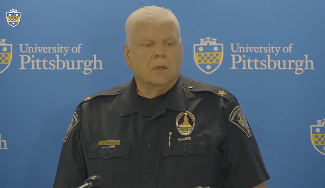 A police chief in a black uniform stands at a podium in front of a blue University of Pittsburgh background