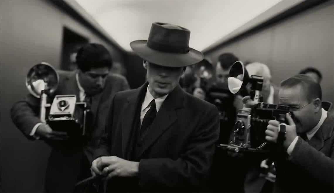 A black and white still of a man being followed with cameras down a hallway from the film "Oppenheimer."